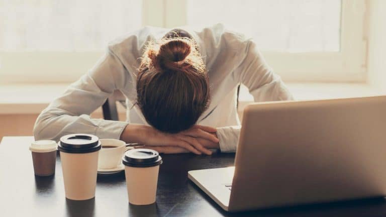 A woman with writer's block is sitting at a desk with her head in her hands. A laptop and three cups of coffee are sitting on the desk in front of her.